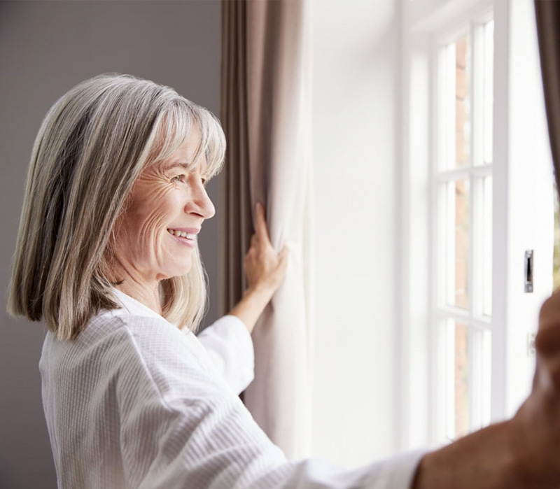 Senior Woman Opening Bedroom Curtains And Looking Out Of Window